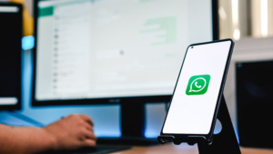 Celular apoiado em suporte com WhatsApp aberto. Pessoa usando WhatsApp Web no PC ao fundo.